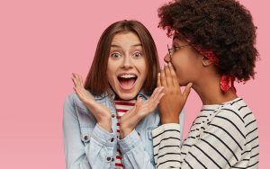 Duas mulheres, uma cliente satisfeita à direita indicando uma empresa para sua amiga, contando com se fosse um segredo, e a outra sorri animada.