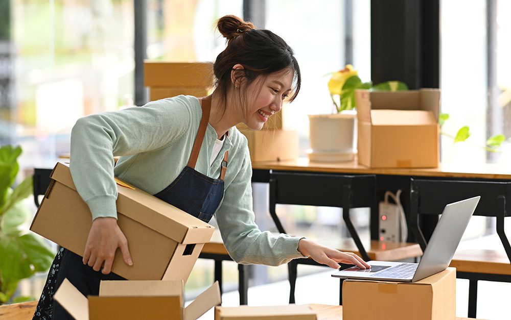 Empreendedora segurando caixa enquanto sorri e confere notebook para verificar pedidos do seu e-commerce